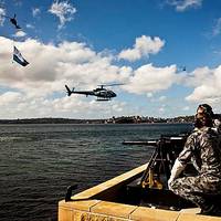 The Royal Australian Navy's Three Pound Saluting Gun Battery prepares to fire across Sydney Harbour as a Squirrel Helicopter decorated with International Fleet Review decals hovers off Garden Island Naval Base as a Seahawk helicopter flying a giant International Fleet Review Flag flys past.